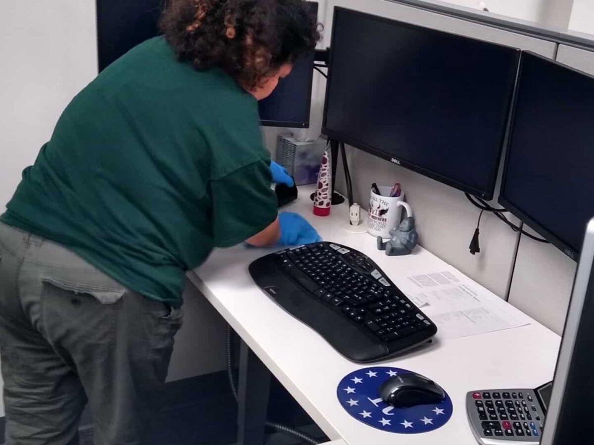 Person cleaning a white desk with dual monitors, keyboard, and office items.