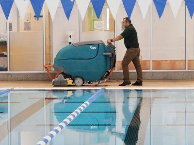 Person pushing a blue pool vacuum across an indoor swimming pool deck.