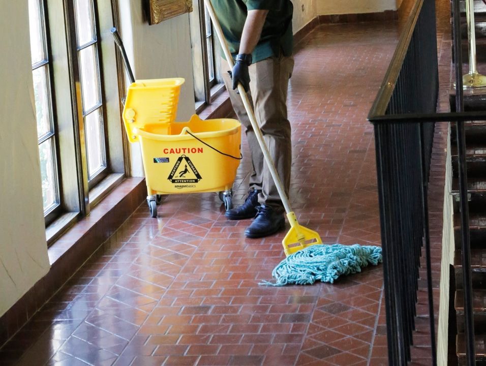 Custodian mopping hallway floor beside yellow caution bucket and cleaning cart