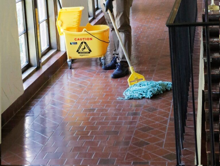 Yellow caution sign and cleaning supplies on a tiled hallway floor, with a mop and bucket by the windows.