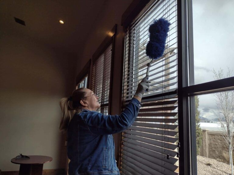 Person dusting window blinds with a blue duster in a sunlit room