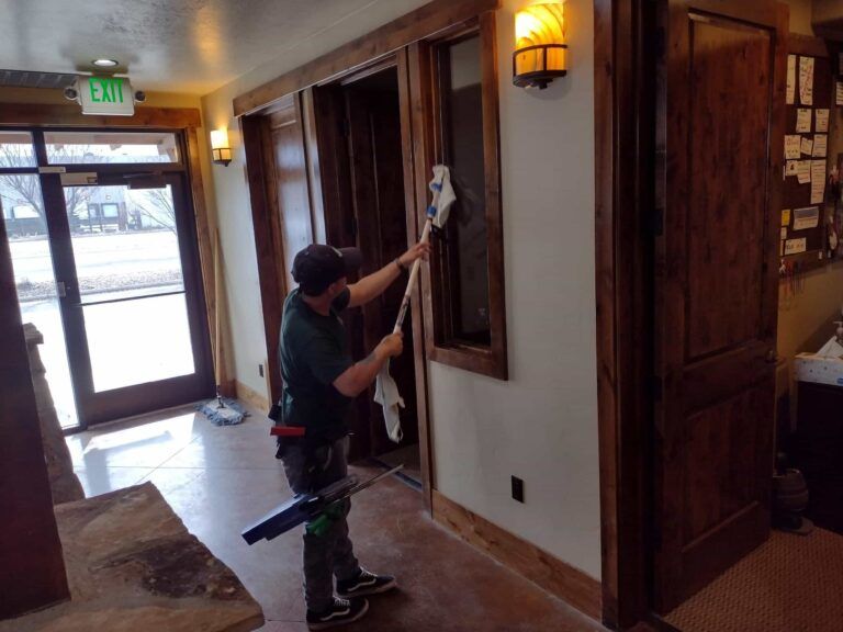 Worker cleaning a window in a wood-paneled hallway near a glass door