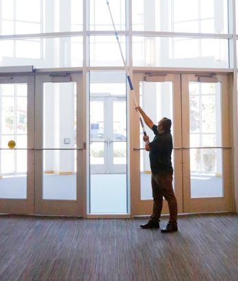 Person using a long pole to adjust a window blind in a bright lobby with glass doors