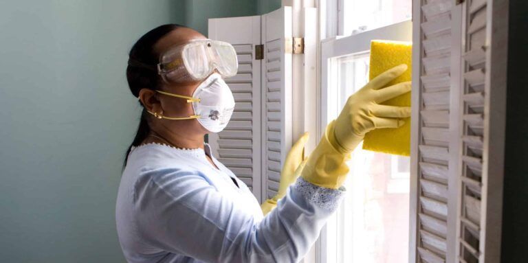 Person wearing gloves and mask cleaning a window with a yellow cloth indoors