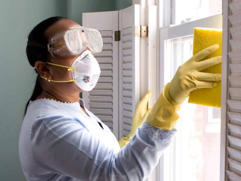Person wearing goggles, mask, and gloves cleaning a window with a yellow sponge