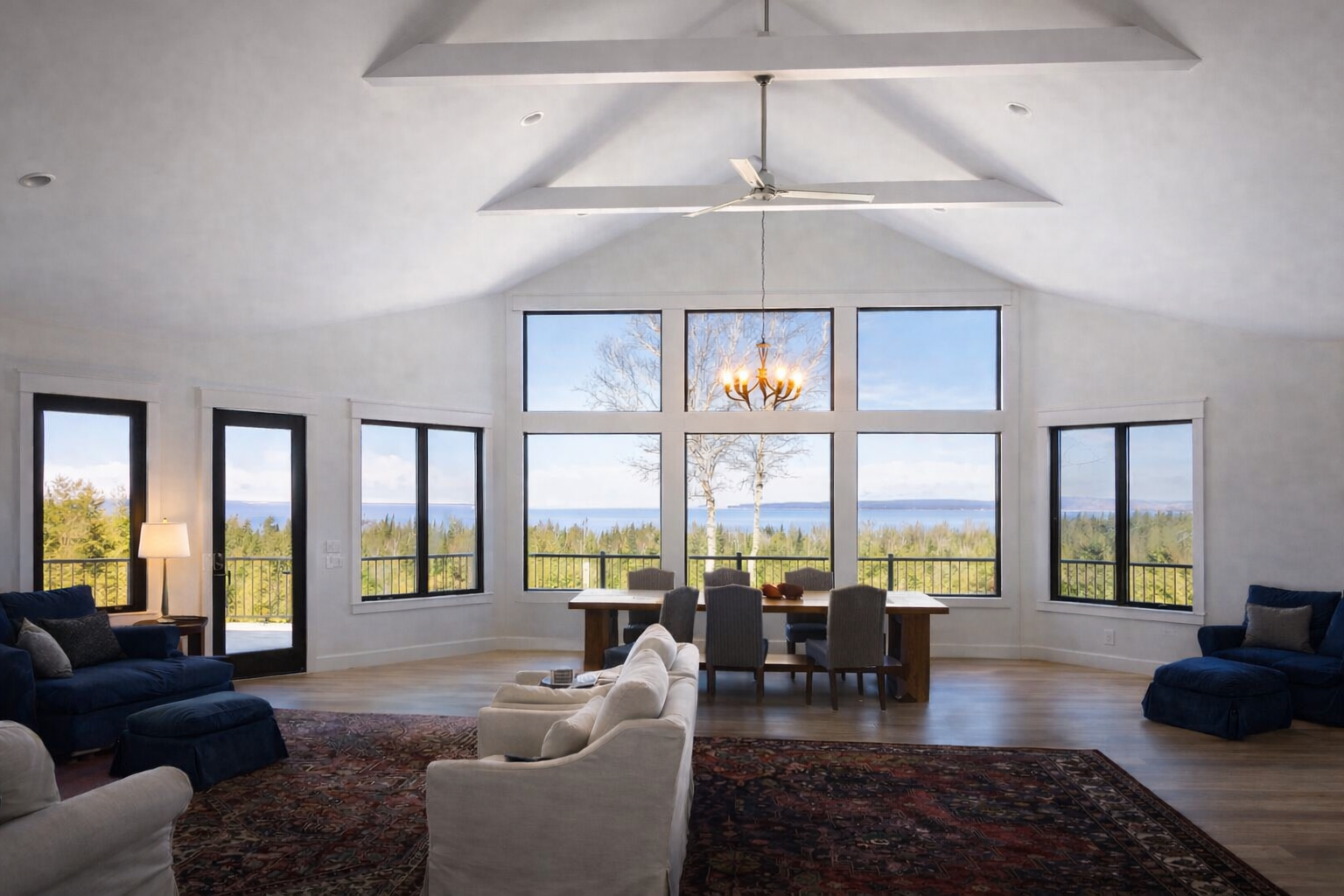 Bright living room with floor-to-ceiling windows, white beams, dark sofas, and a dining table by the view