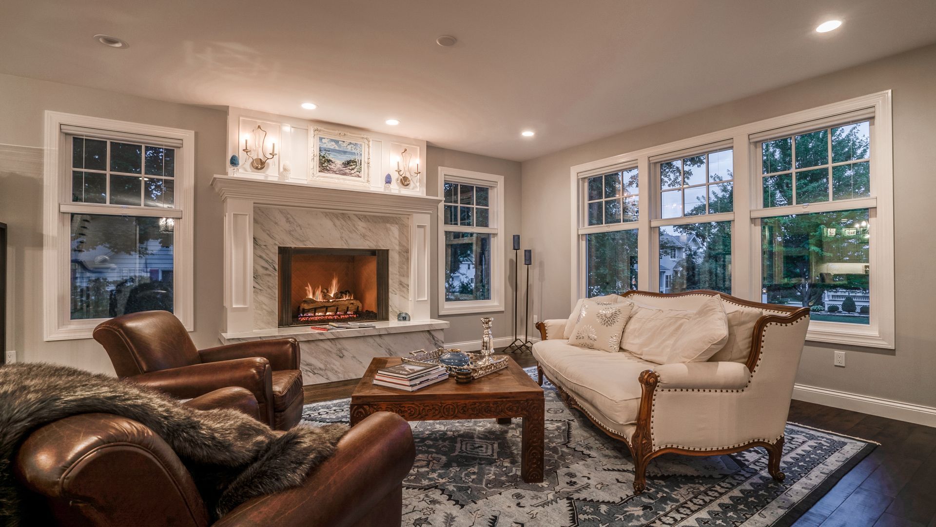Bright living room with a fireplace, leather chairs, a cream sofa, and large windows at dusk