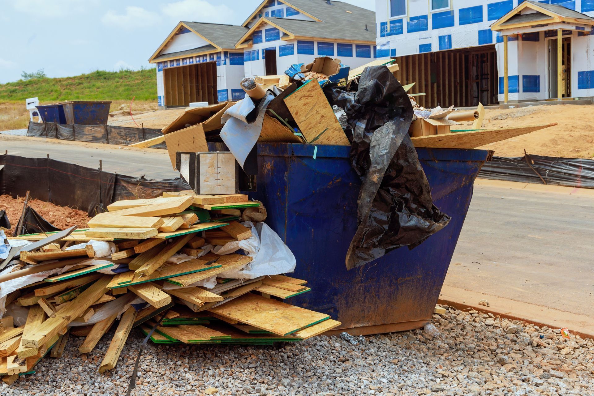 A blue construction dumpster overflowing with wood scraps and debris sits on a gravel road in front of unfinished houses.