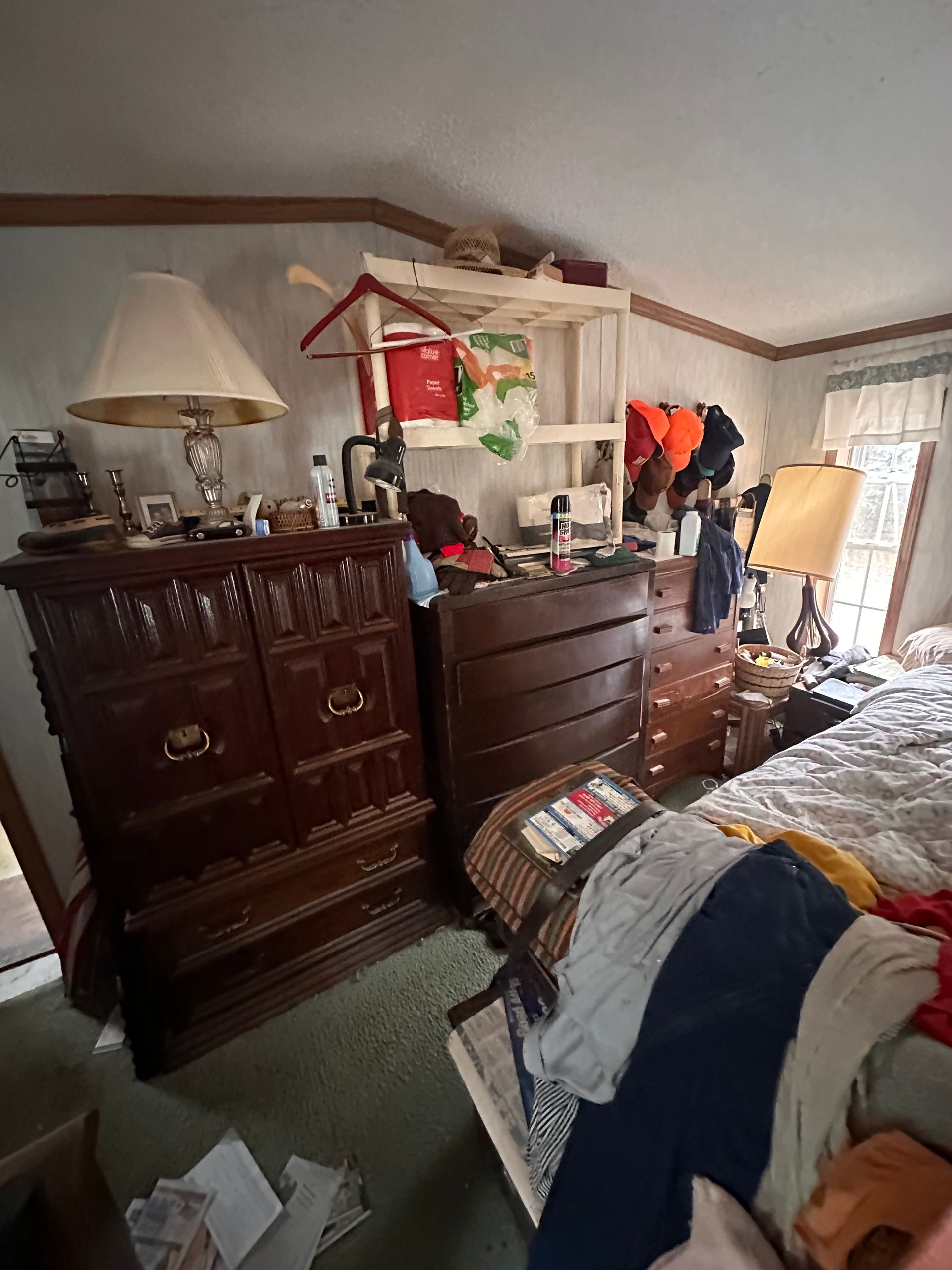 A bedroom with dark wood dressers, a lamp, a wooden shelving unit with clothes, and a bed covered in scattered clothing.