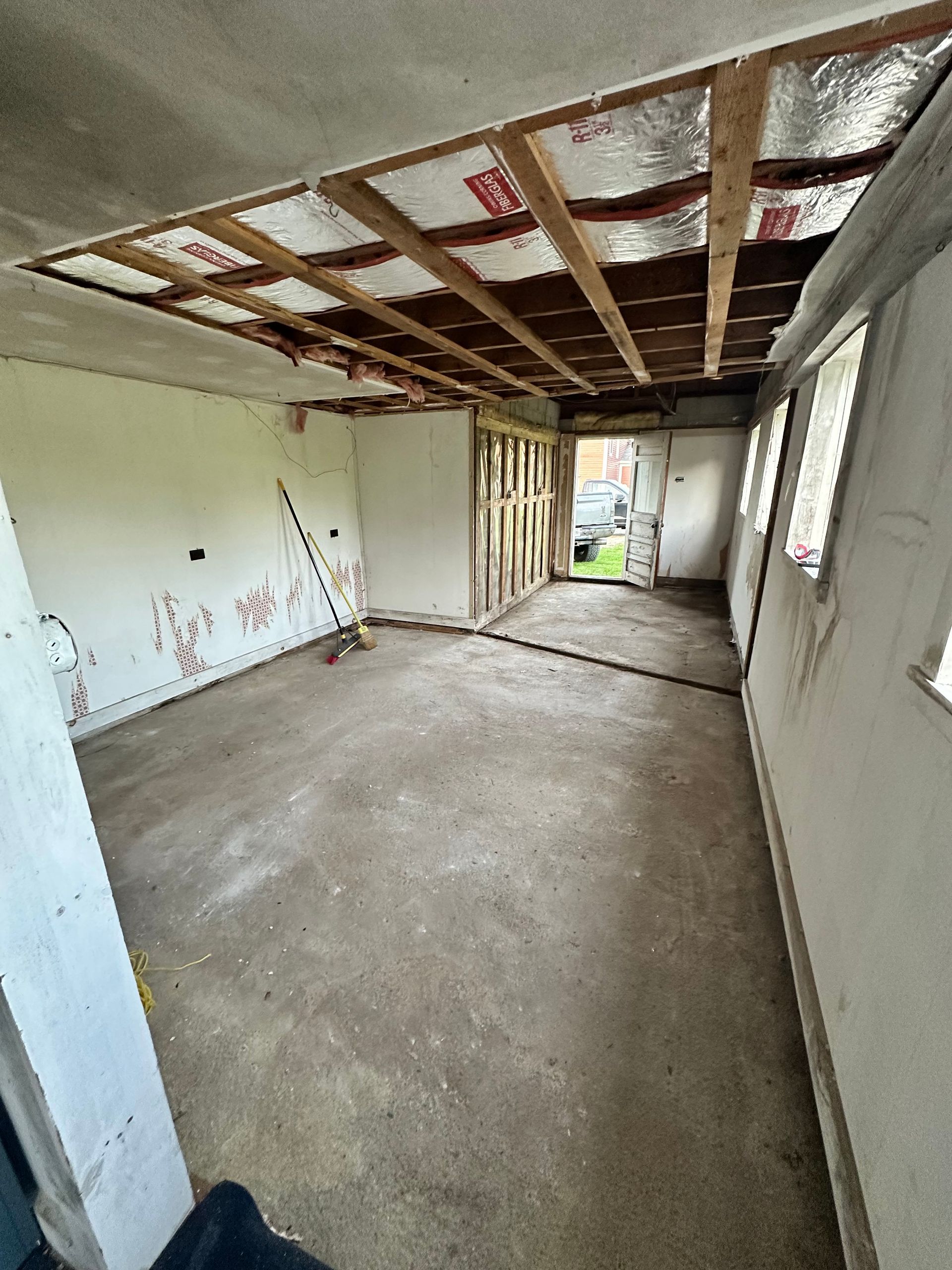 A room under renovation with an exposed ceiling frame, unpainted white walls, concrete floor, and windows to the right.