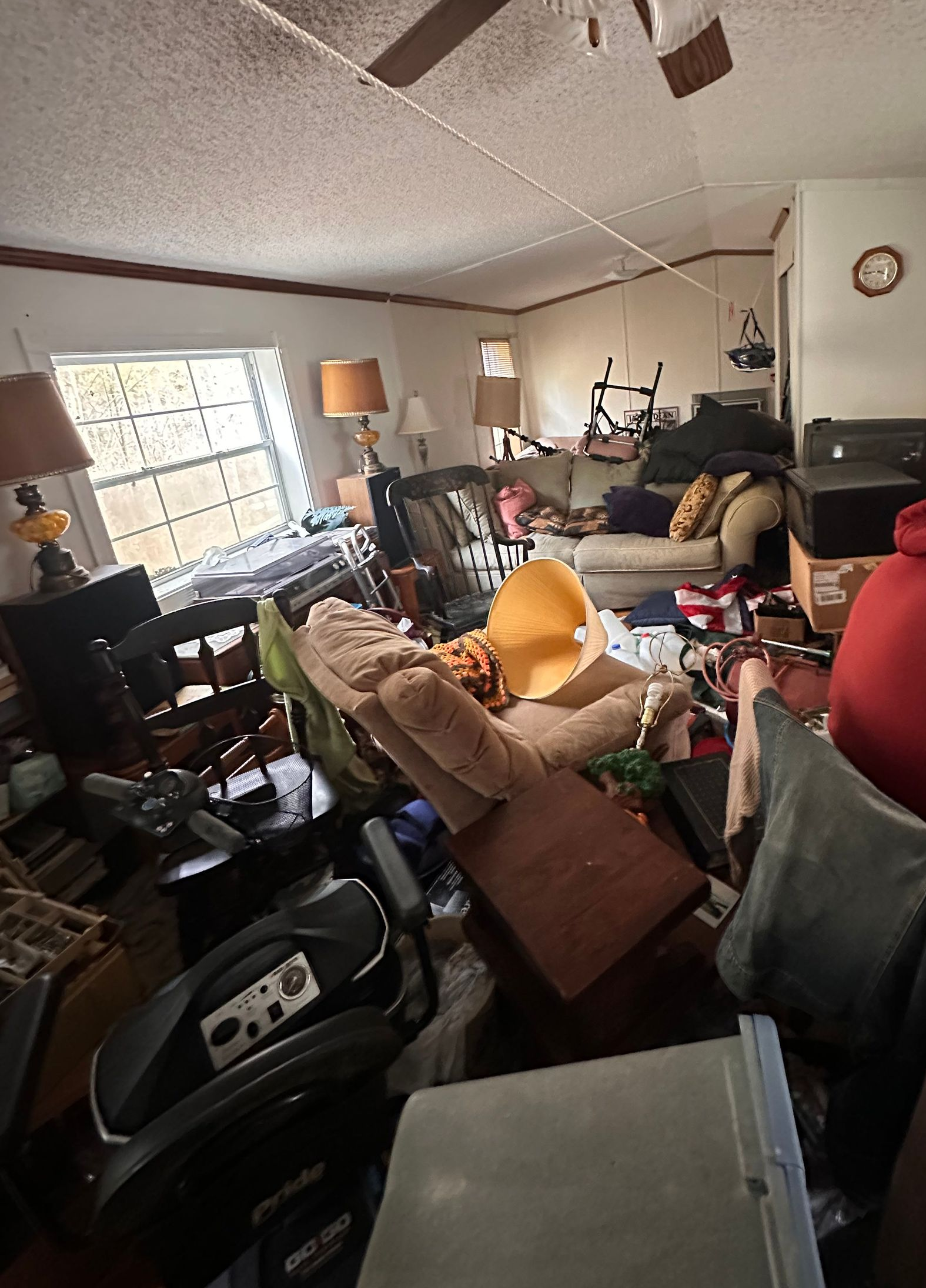 A man in a red hoodie sits in a cluttered living room filled with furniture, boxes, and various household items.