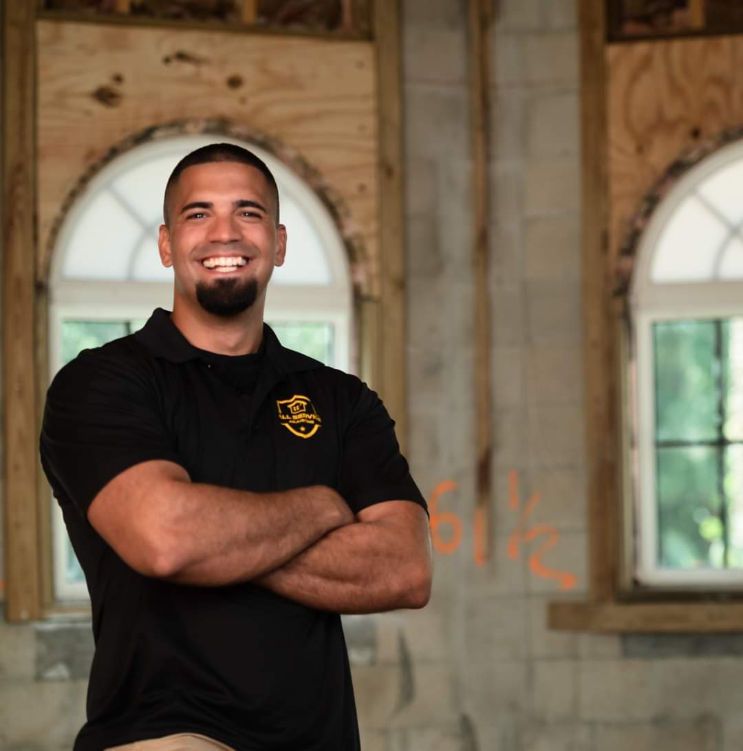 A man wearing a black shirt with a shield on it smiles with his arms crossed