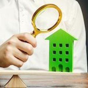 A person is holding a magnifying glass over a green house on a scale.