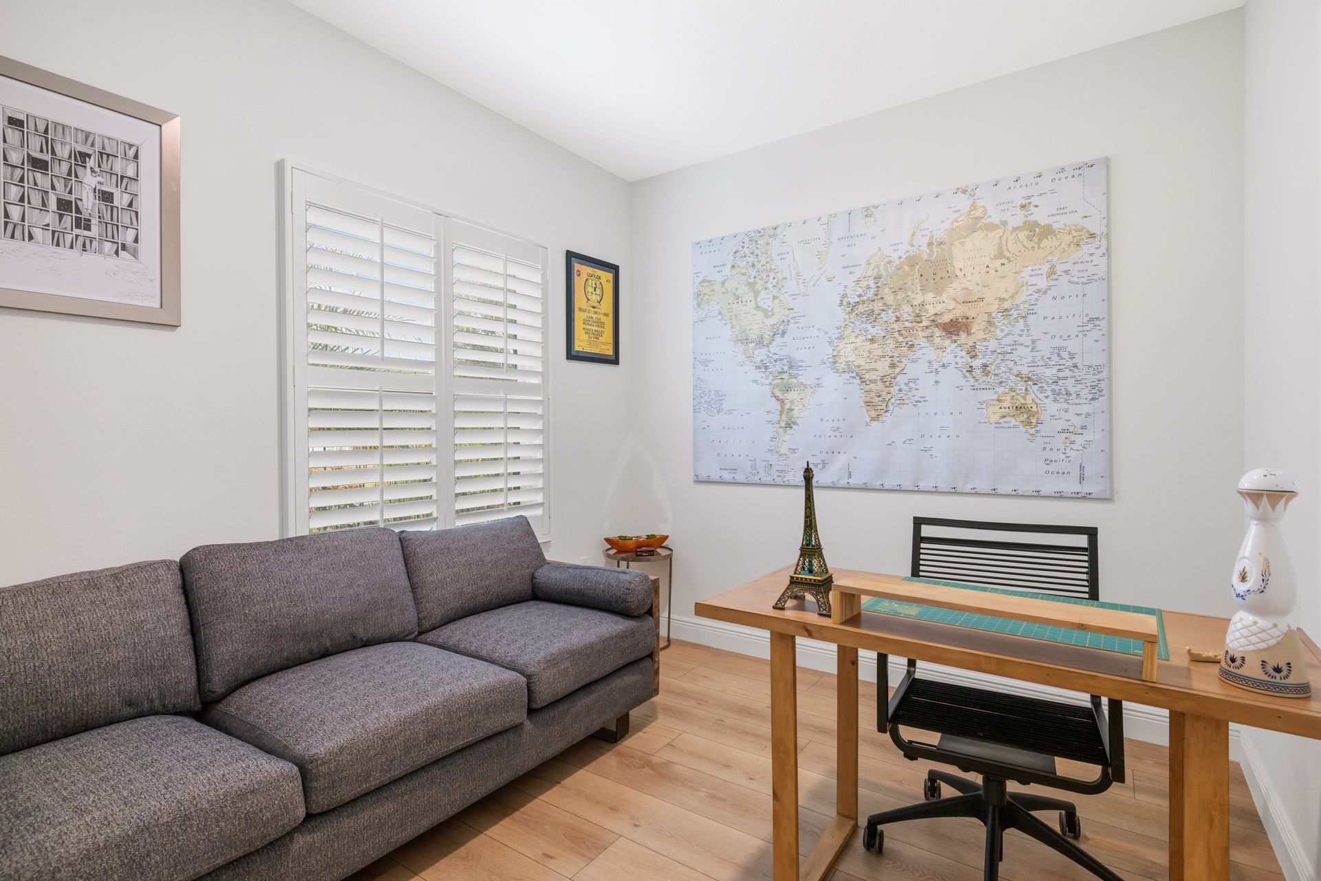 A home office featuring a gray sofa, a wood desk with a black rolling chair, and a large world map on the wall.