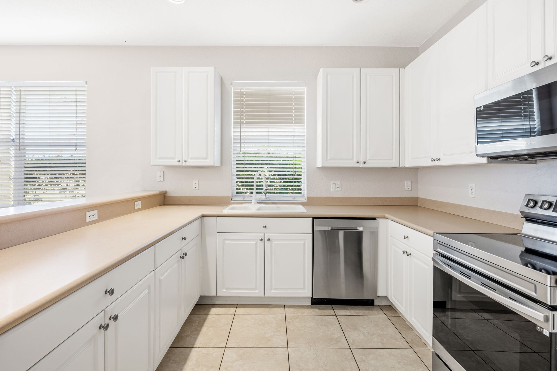 A bright kitchen with white cabinets, beige countertops, stainless steel appliances, and tiled floors.