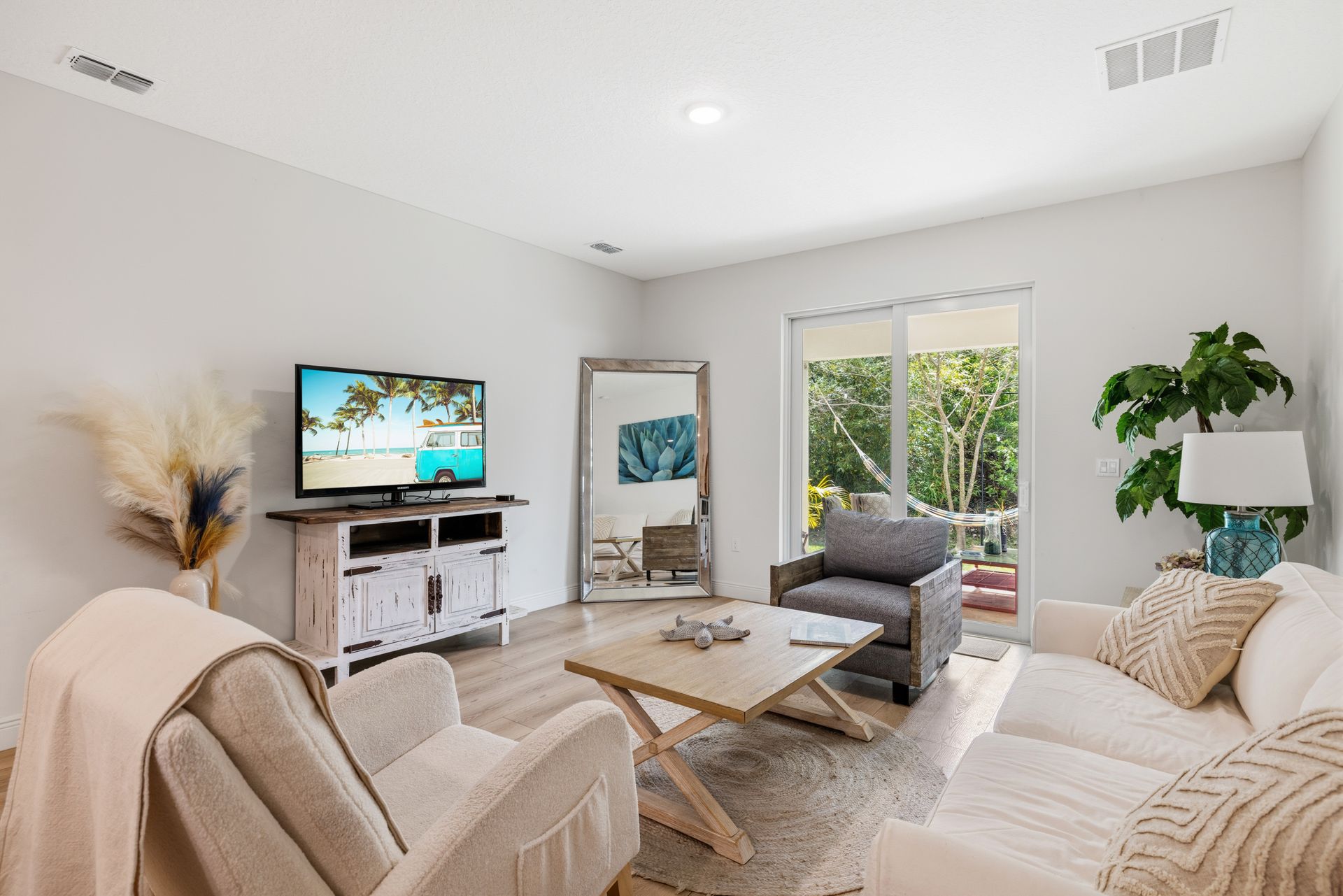 A living room with light walls, a beige sofa and armchair, a wooden coffee table, a TV on a stand, and a floor mirror.