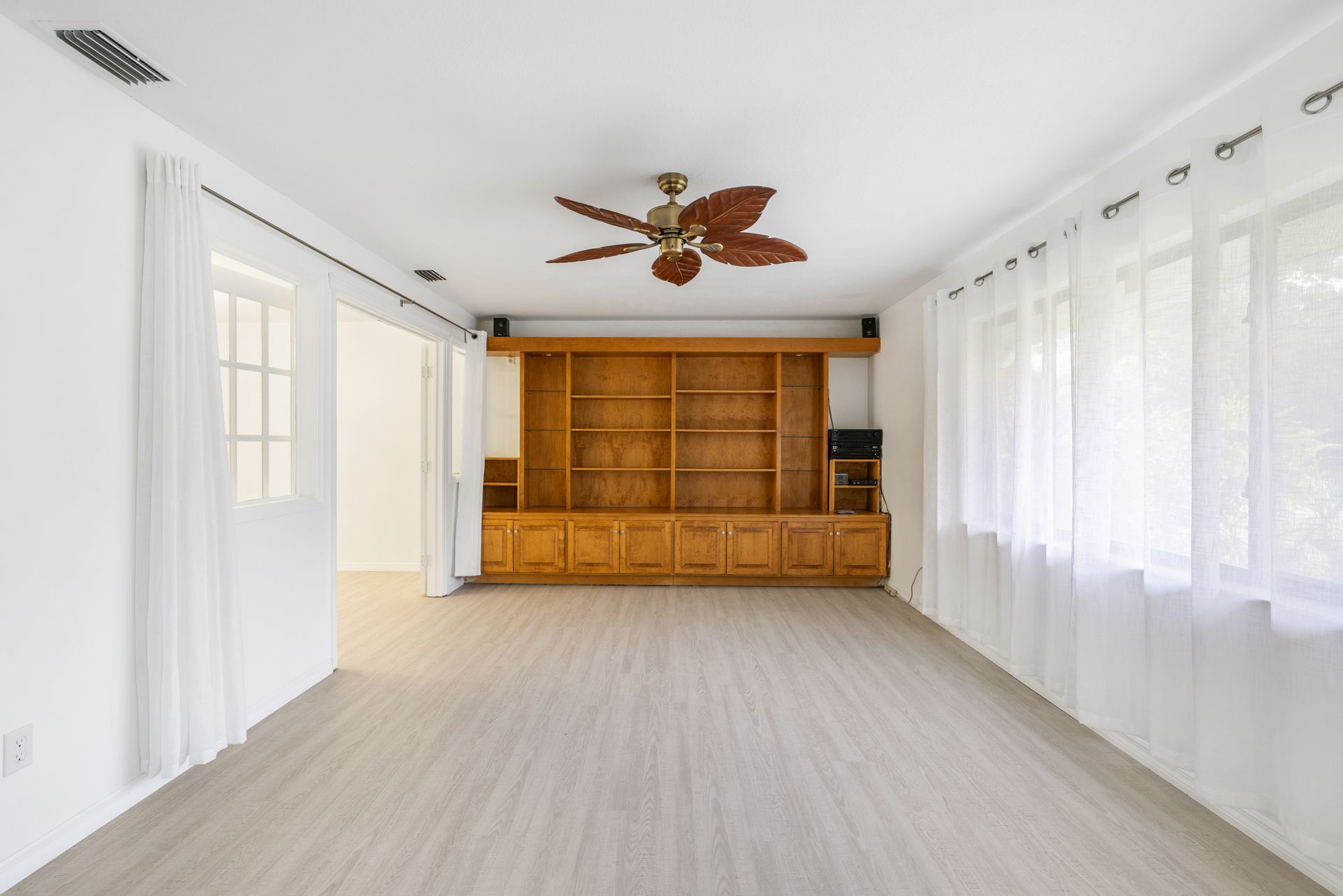 A bright, empty living room with light wood-style flooring, white walls, a wooden bookshelf, and a ceiling fan.