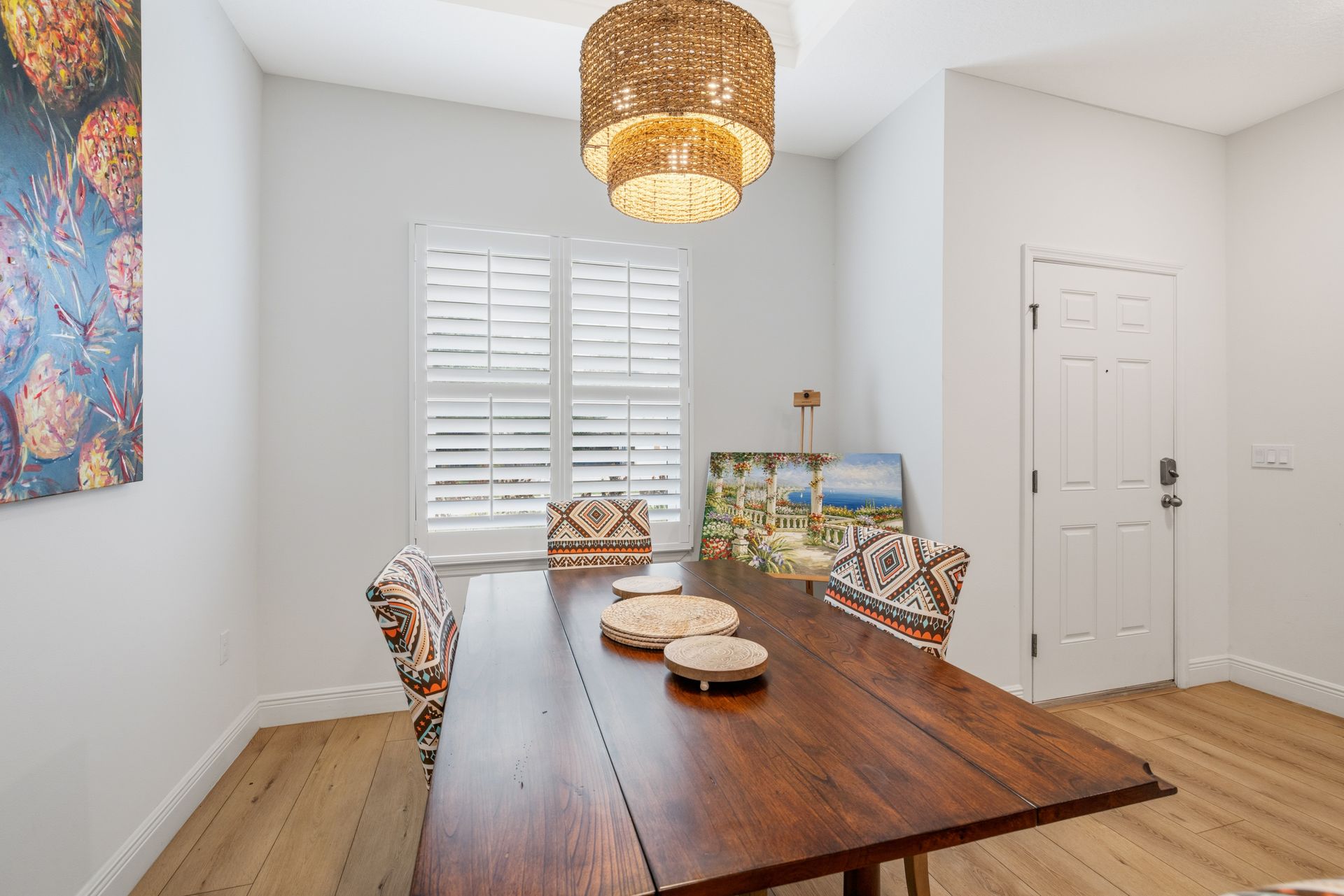 Dining room featuring a wooden table, patterned chairs, and a rattan chandelier, with a window and a white door.