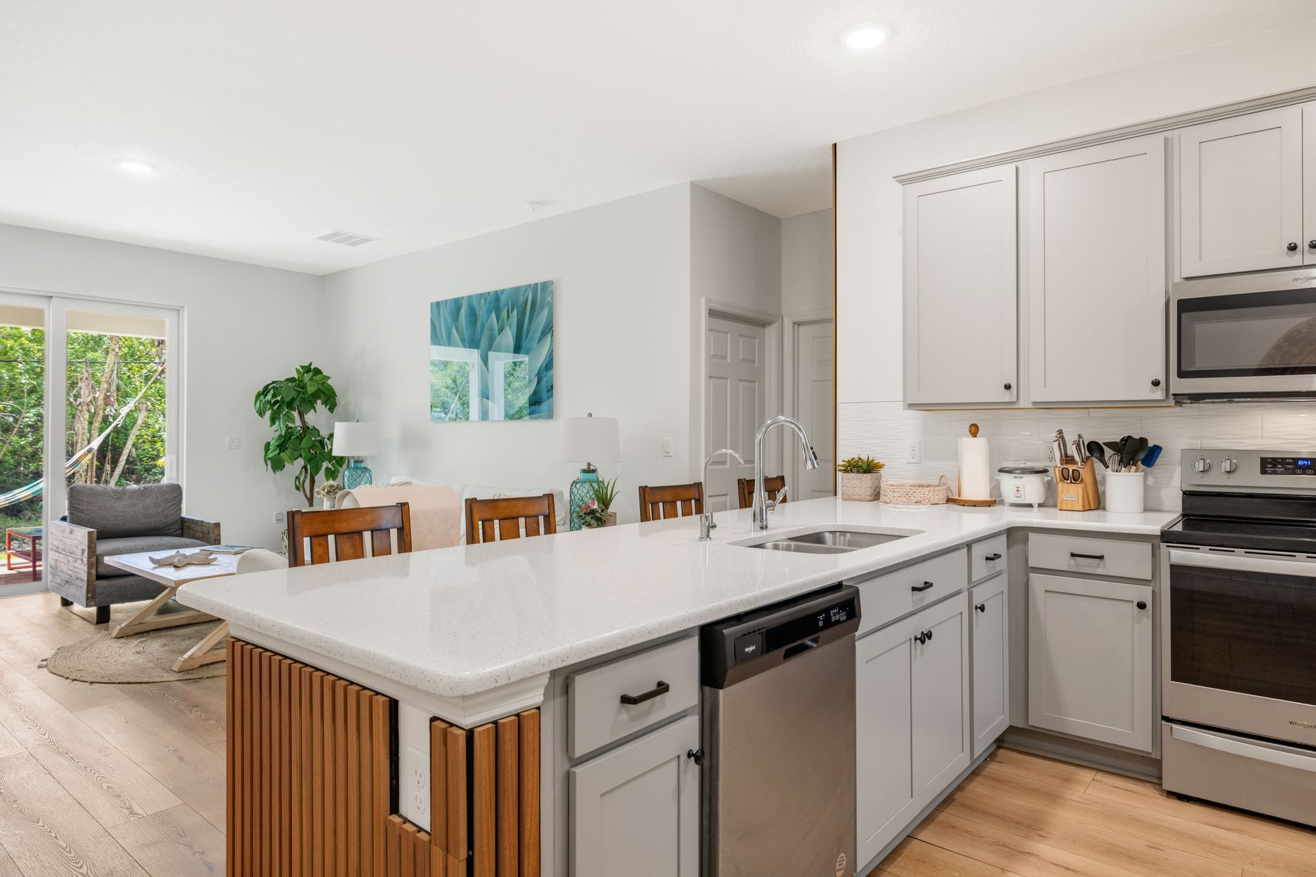 Modern kitchen with light gray cabinets, a white island, stainless steel appliances, and a view into a living room.