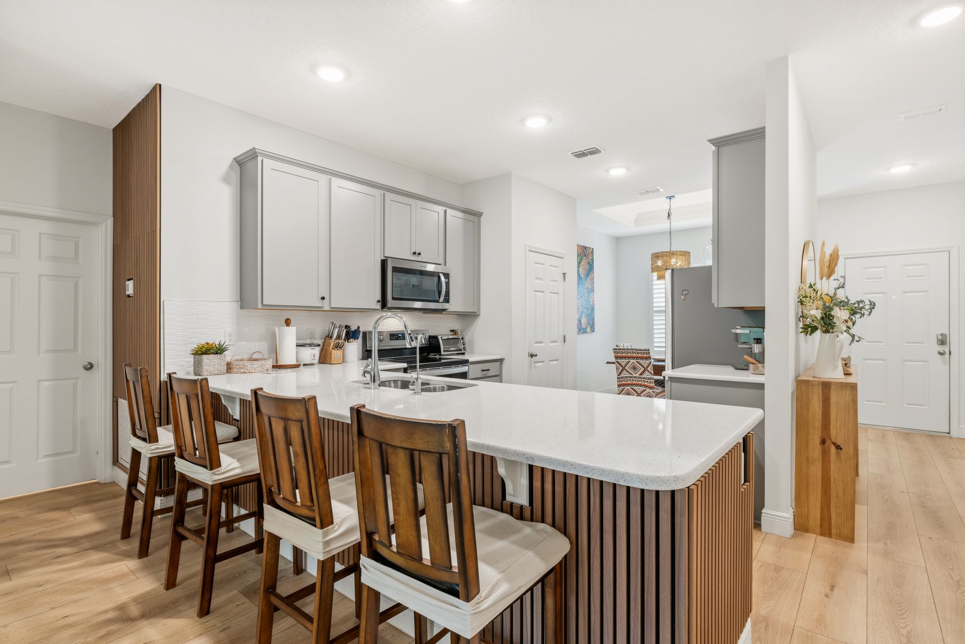 Modern kitchen with gray cabinets, white countertops, four wooden bar stools, and light wood floors.