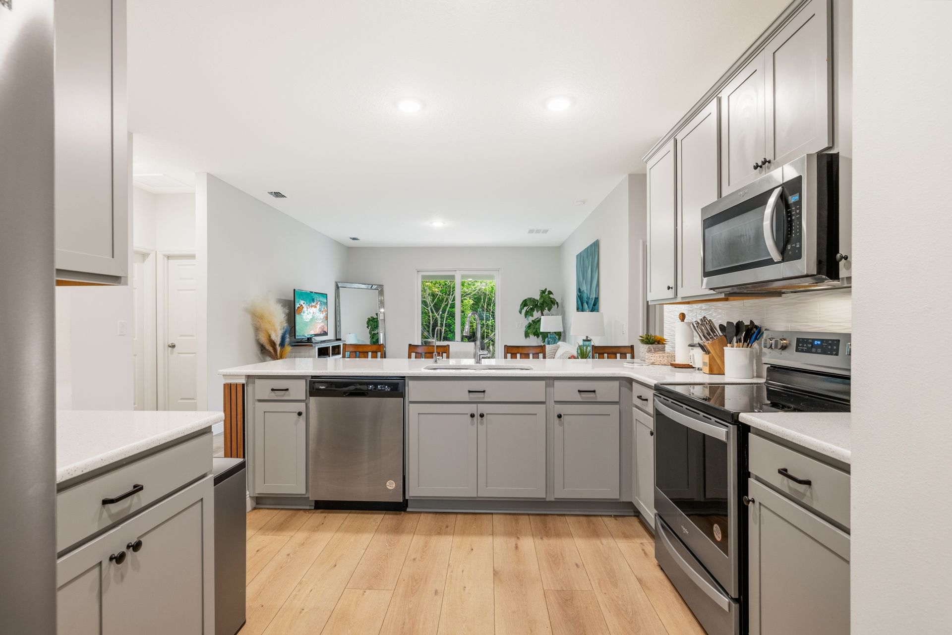 A modern, well-lit kitchen with light gray cabinets, stainless steel appliances, and wood floors, opening to a dining area.