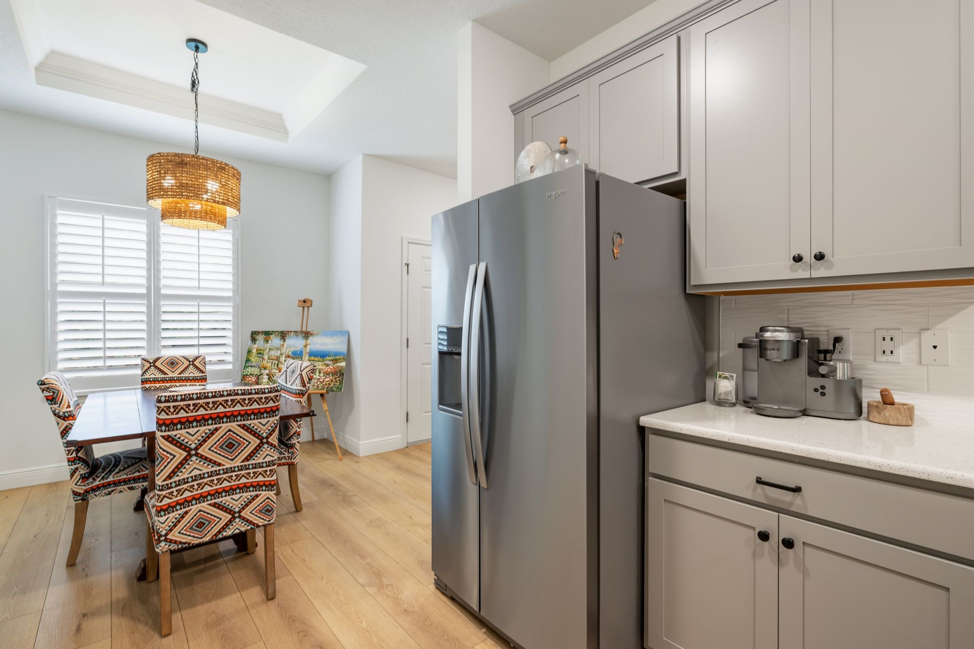 A kitchen with gray cabinets, a stainless steel refrigerator, and a small dining area with patterned chairs.