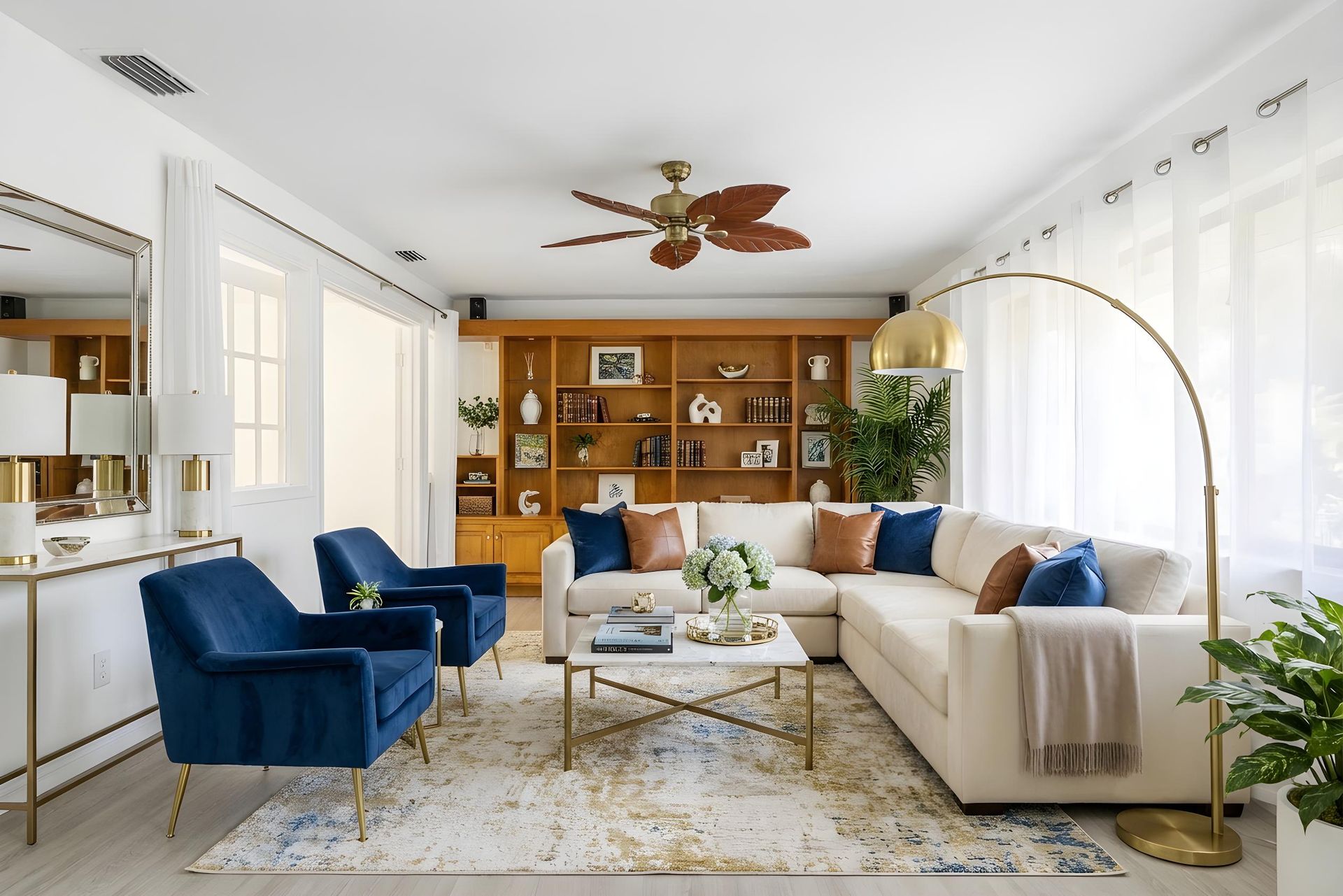 A bright living room features a cream sectional sofa, two blue armchairs, a marble coffee table, and a large wood bookshelf.