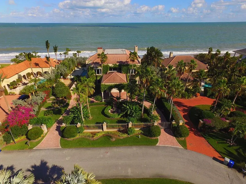 Aerial view of luxury homes on a beach, lush green landscaping, blue ocean, and a sunny sky.