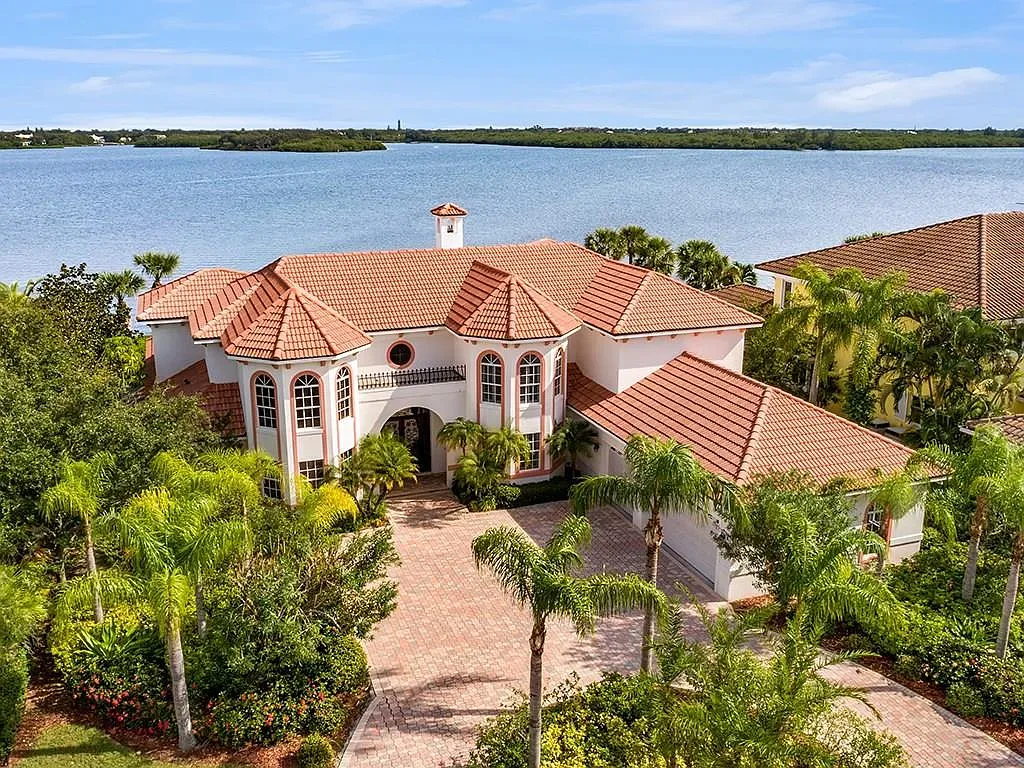 A large, white Mediterranean-style house with a terracotta tile roof, set among lush tropical trees overlooking a bay.