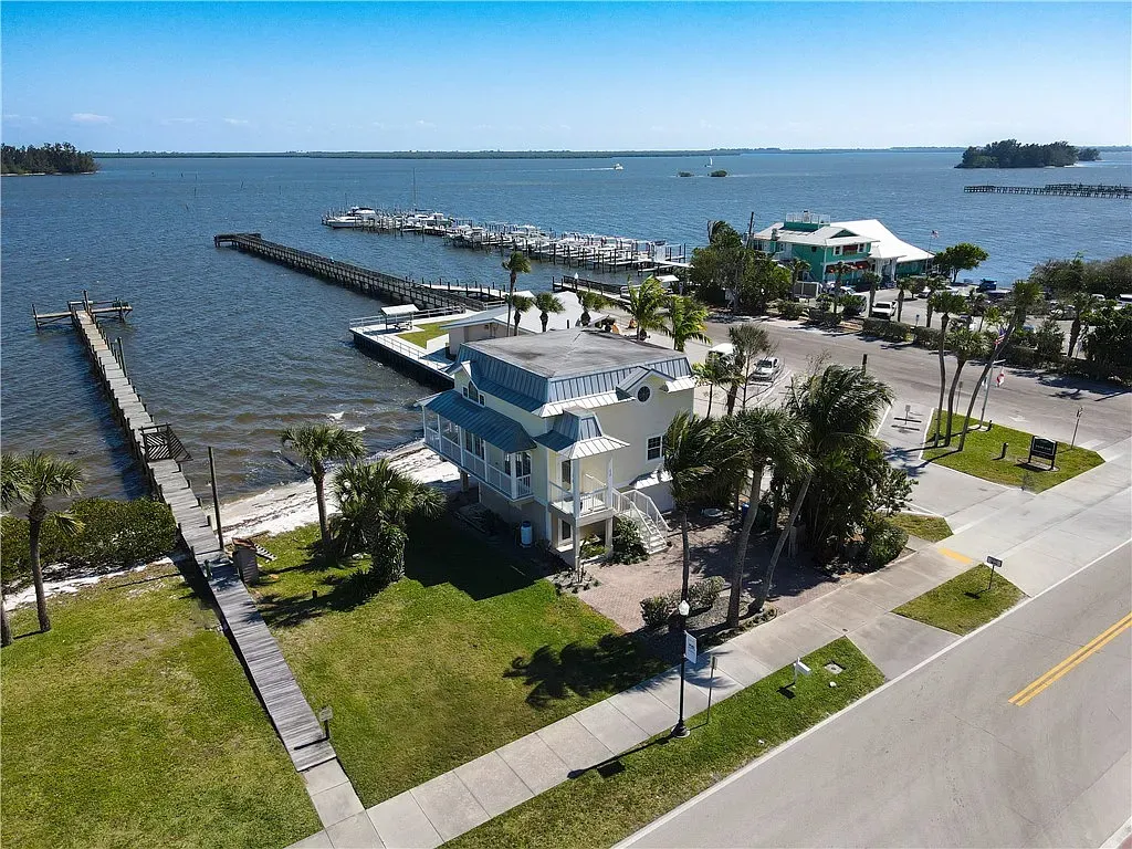 An aerial view of a yellow waterfront house with a long wooden pier and a marina filled with boats under a clear blue sky.