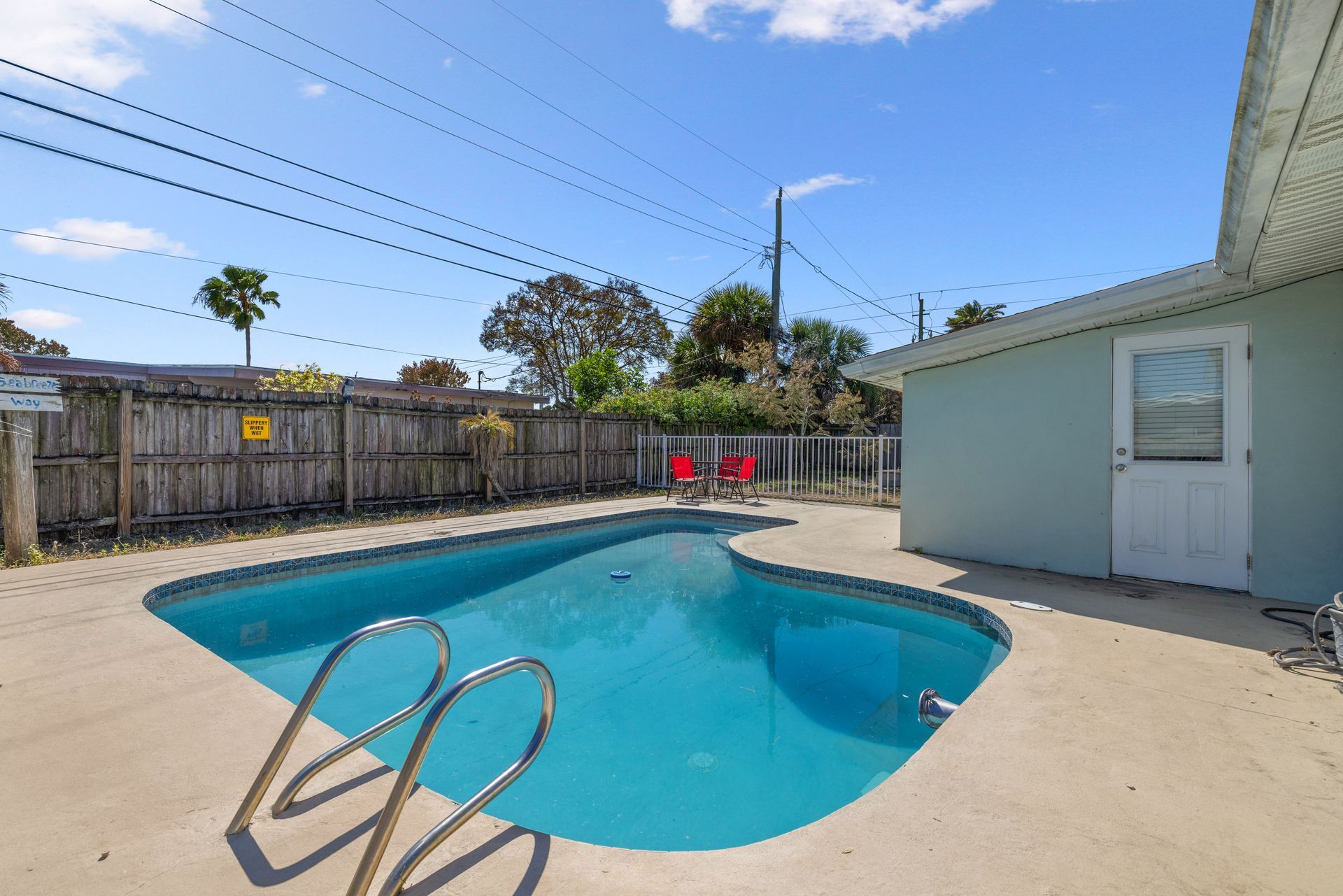 A light blue swimming pool in a sunny backyard with a concrete patio, wooden fence, and part of a house.