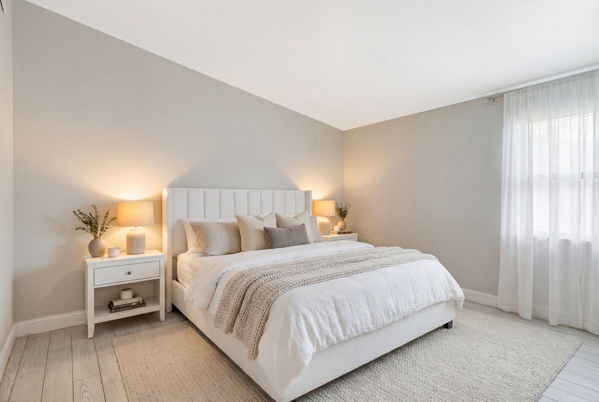 A bright, neutral-toned bedroom with a white upholstered bed, side tables with lamps, and sheer curtains over a window.