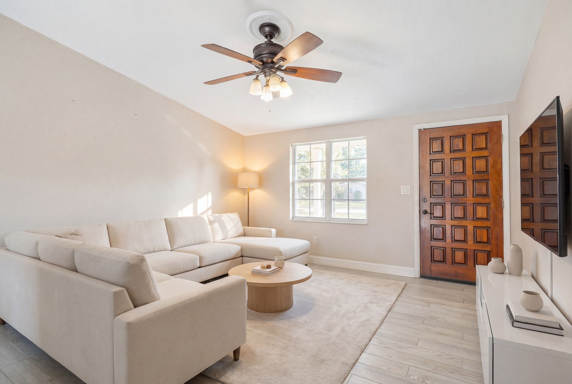 A bright, modern living room featuring a large beige sectional sofa, a round coffee table, a wood door, and a ceiling fan.