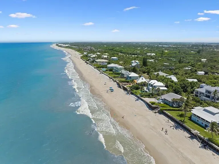 Aerial view of a sandy beach bordered by houses and dense green trees along the ocean coastline under a clear blue sky.