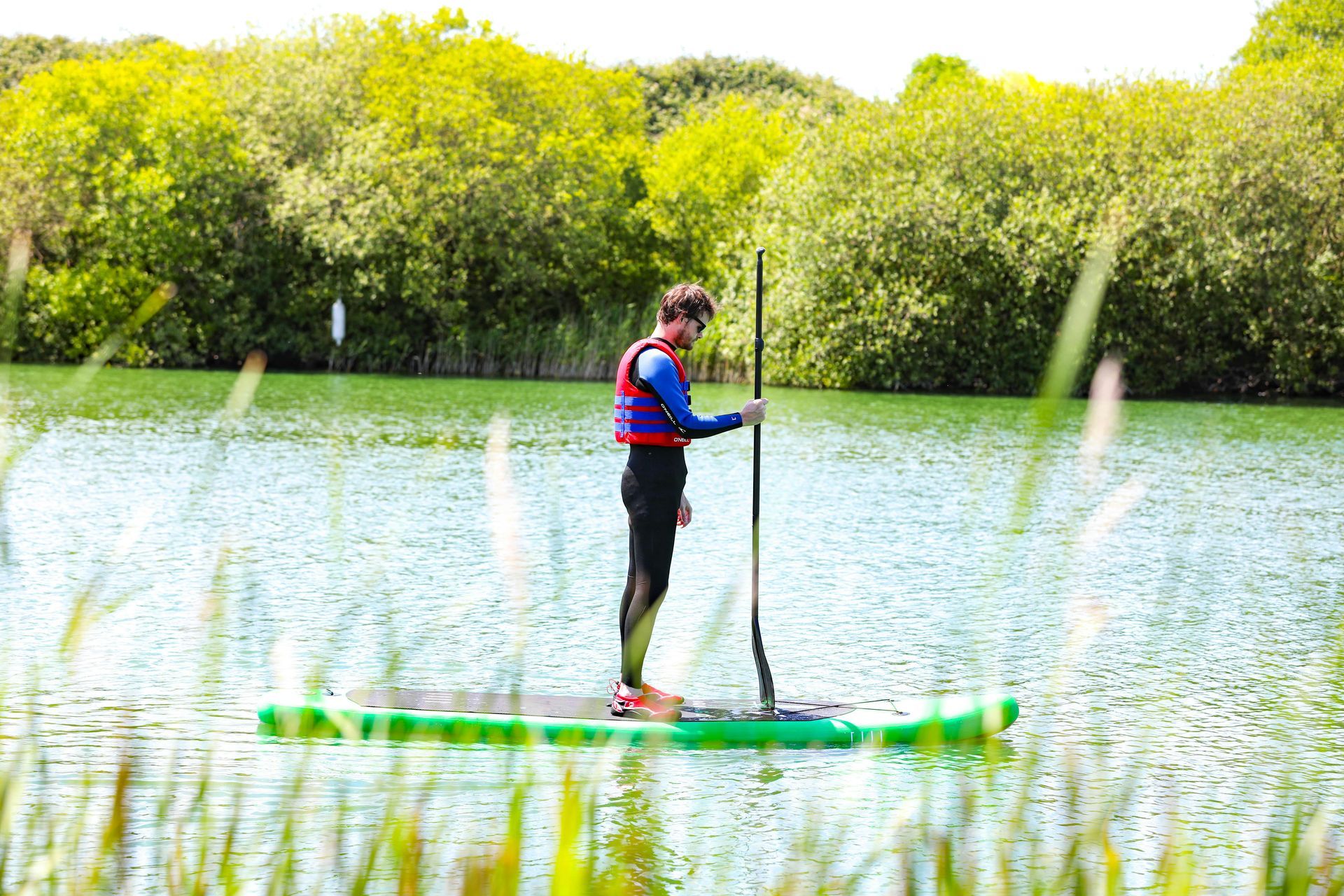 A person in a life jacket stands on a green paddleboard on a calm lake, holding a paddle against a backdrop of trees.