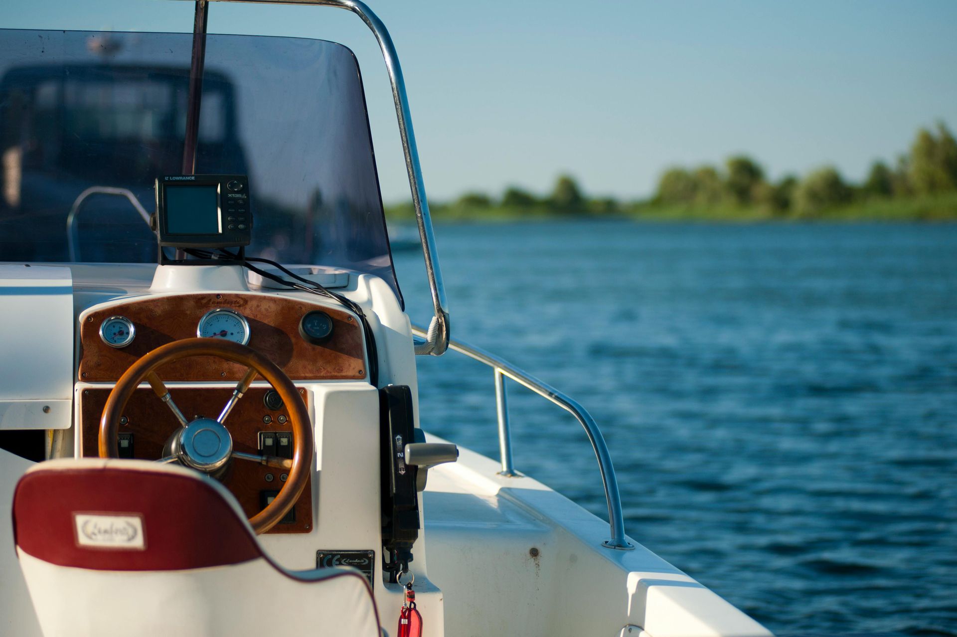 Steering wheel and dashboard of a white motorboat on a blue lake with trees in the background.