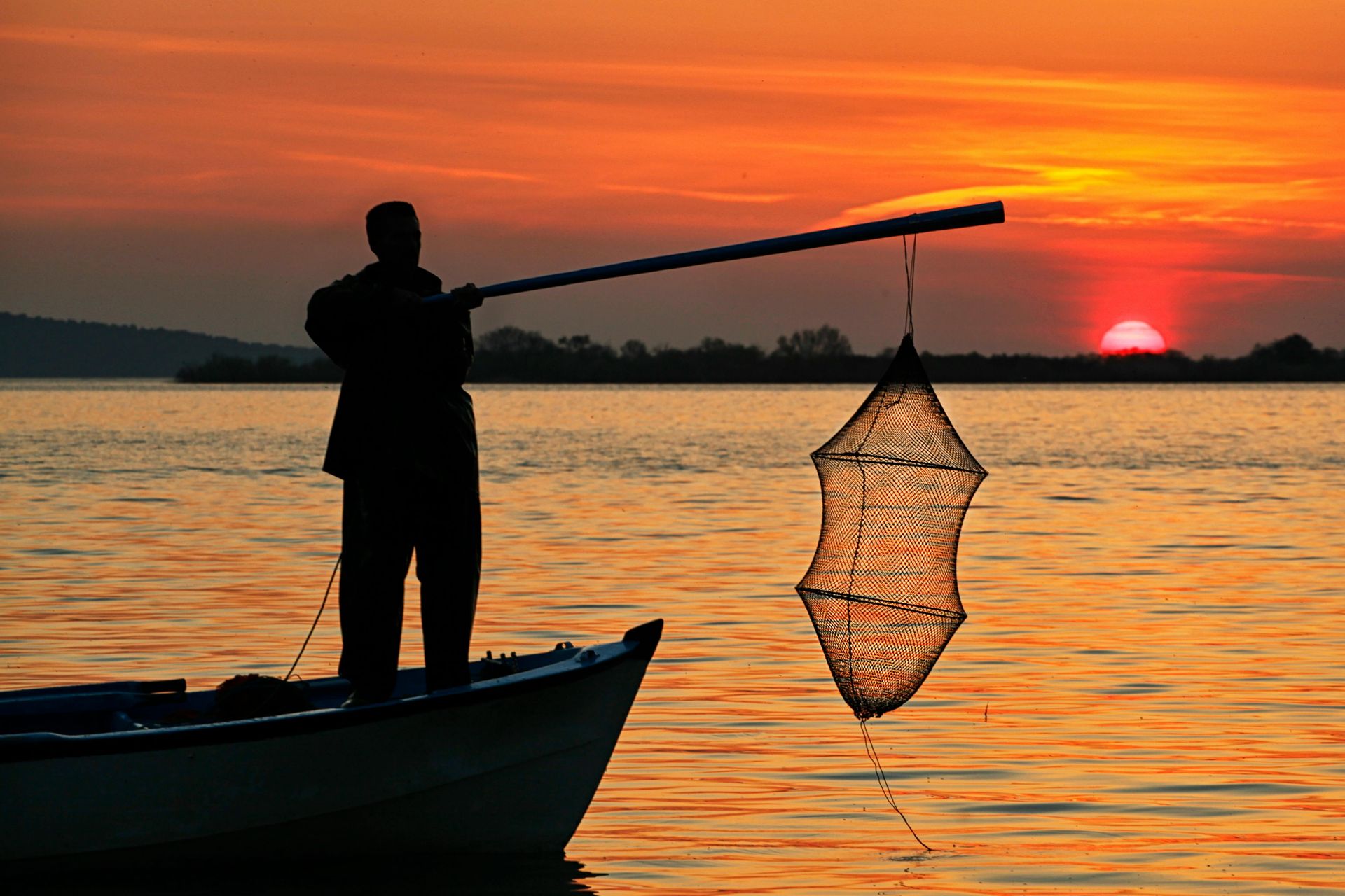 Silhouette of person fishing from a boat at sunset, using a net. Orange sky over water.