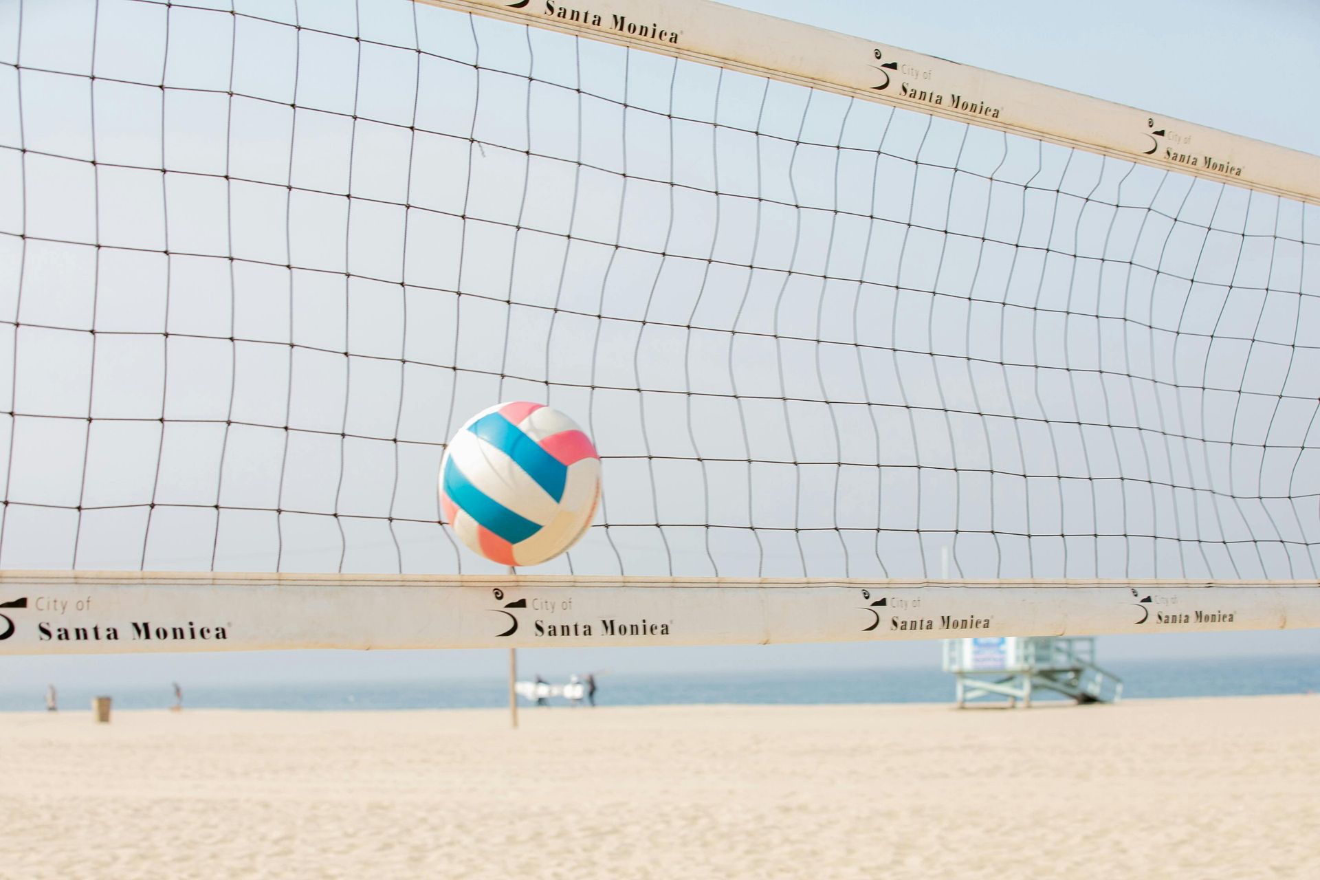 Volleyball in mid-air, near a net on a sandy beach.  Santa Monica in the background.