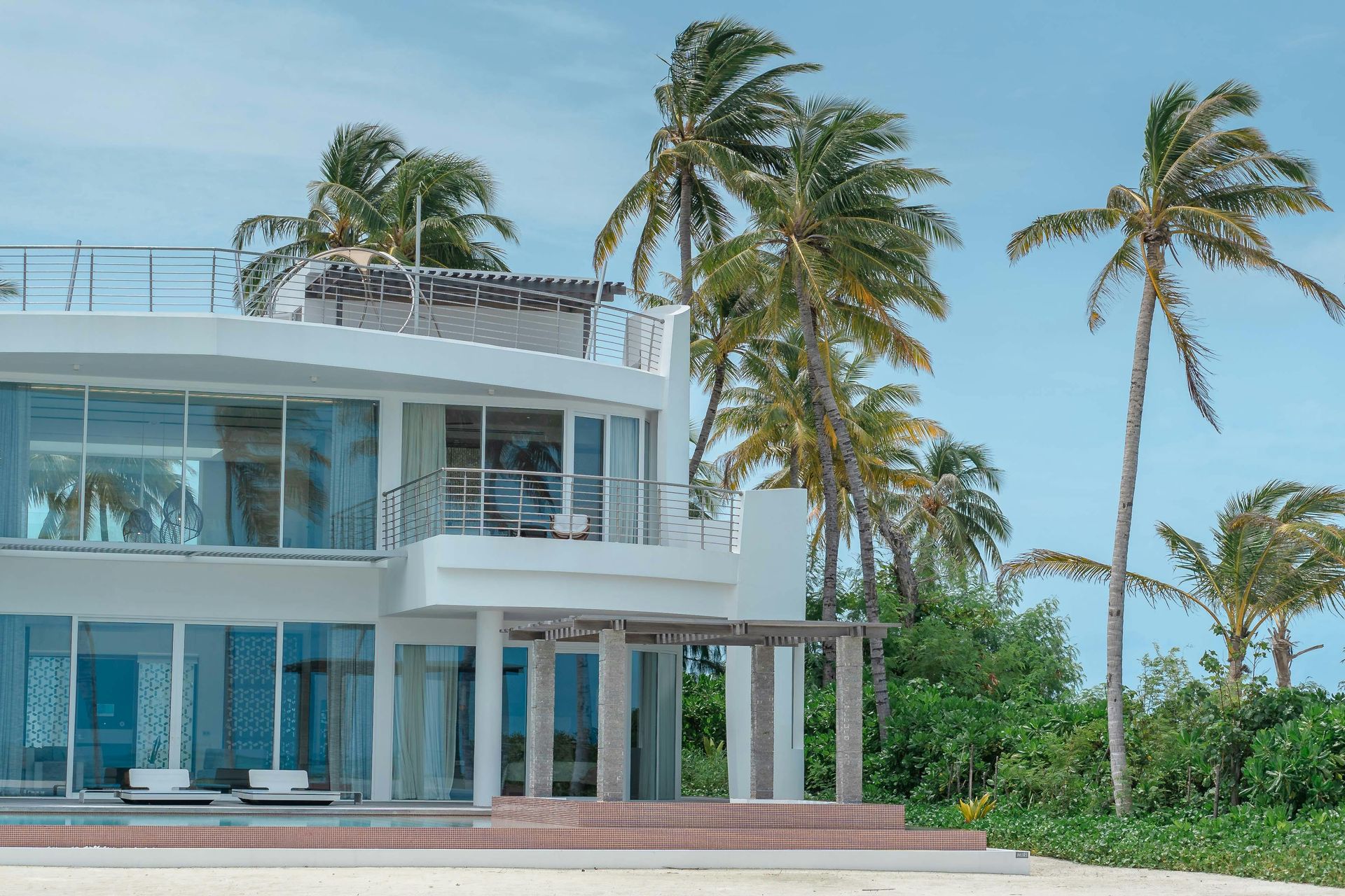 Modern white beachfront villa with palm trees and a blue sky.