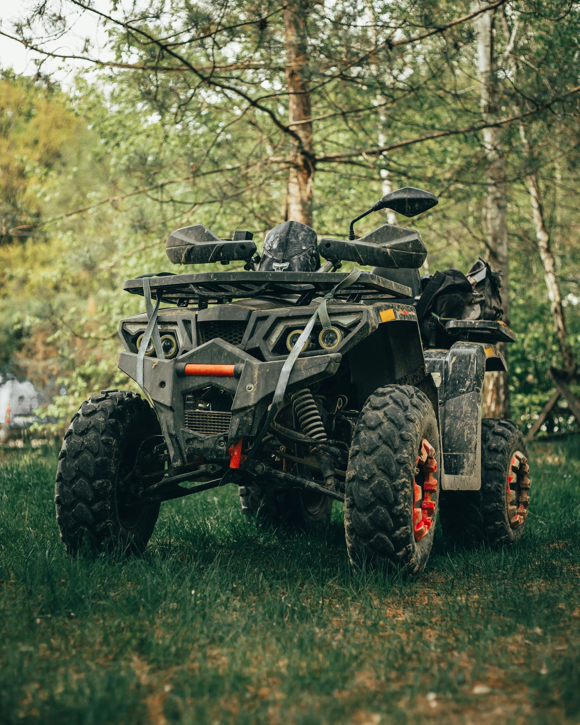 A black all-terrain vehicle parked on a grassy forest floor, featuring orange accents on the wheels and front bumper.