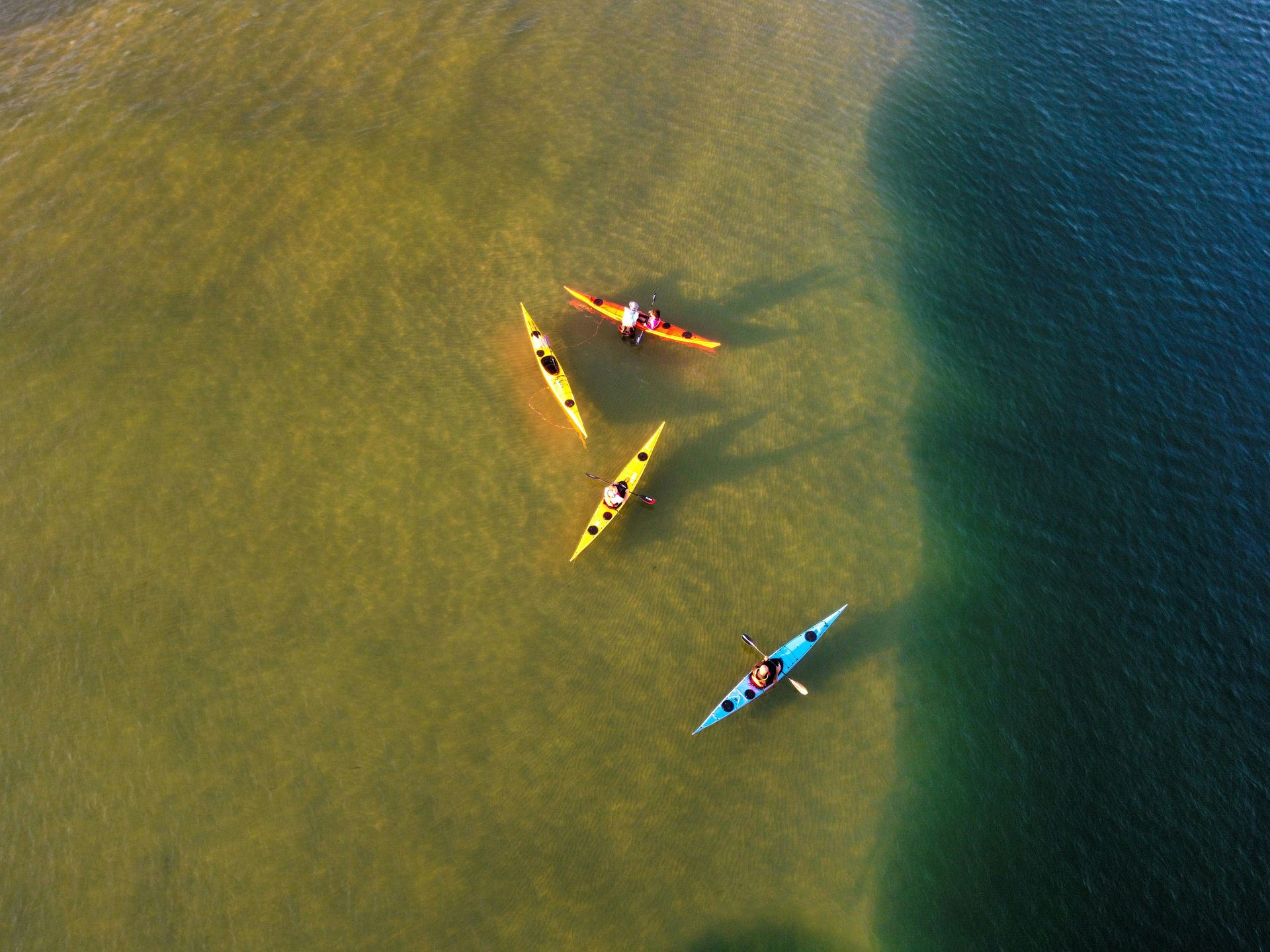 Three kayaks on water, each with a person paddling. Water shades from tan to blue.