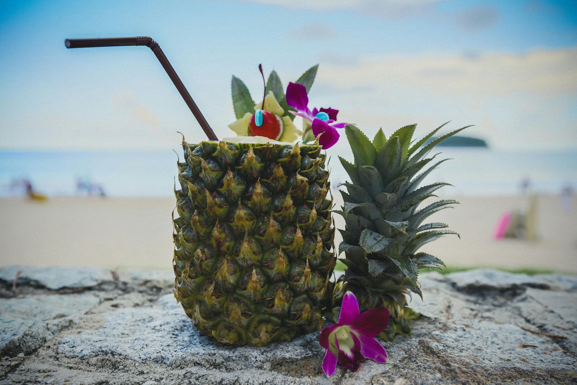 Pineapple cocktail with straw, flowers, and beach background.