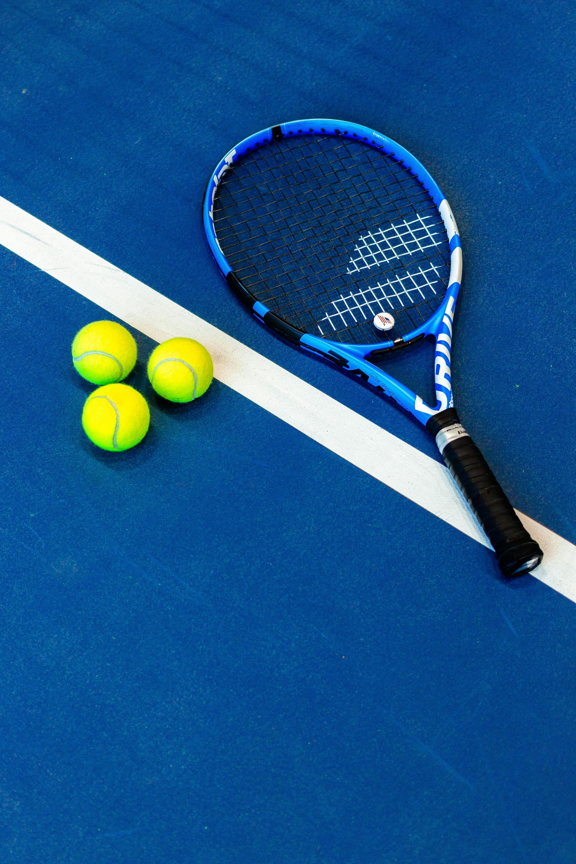 Blue tennis racket and three yellow tennis balls on a blue court next to a white line.