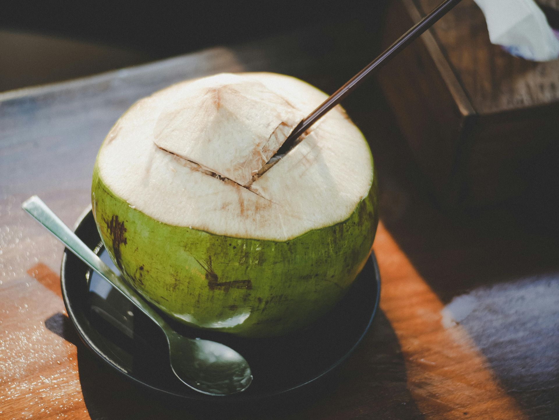 Fresh coconut with straw and spoon on a small plate, on a wooden surface.