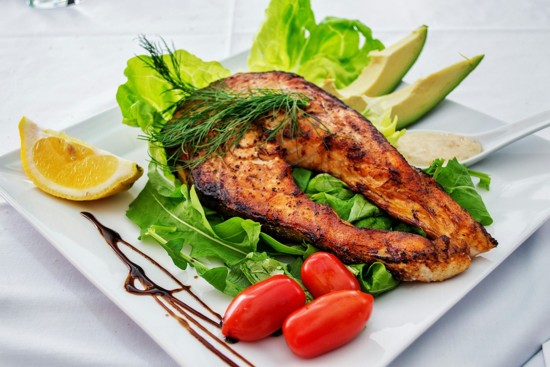 Grilled salmon steak on a white plate with lettuce, avocado, tomatoes, lemon, and sauce.