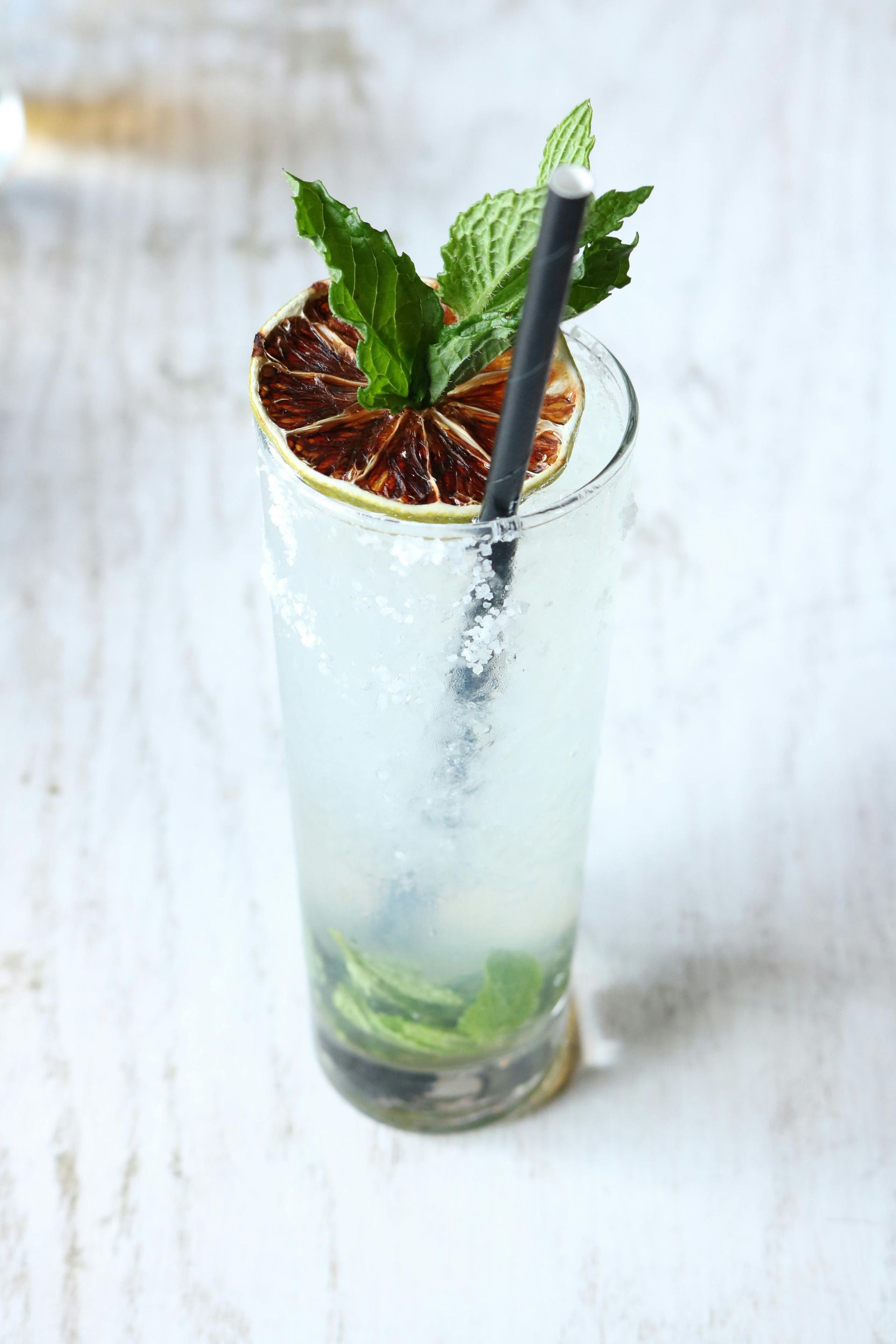 Tall cocktail with lime garnish, mint sprig, black straw, in a clear glass on a white wooden surface.
