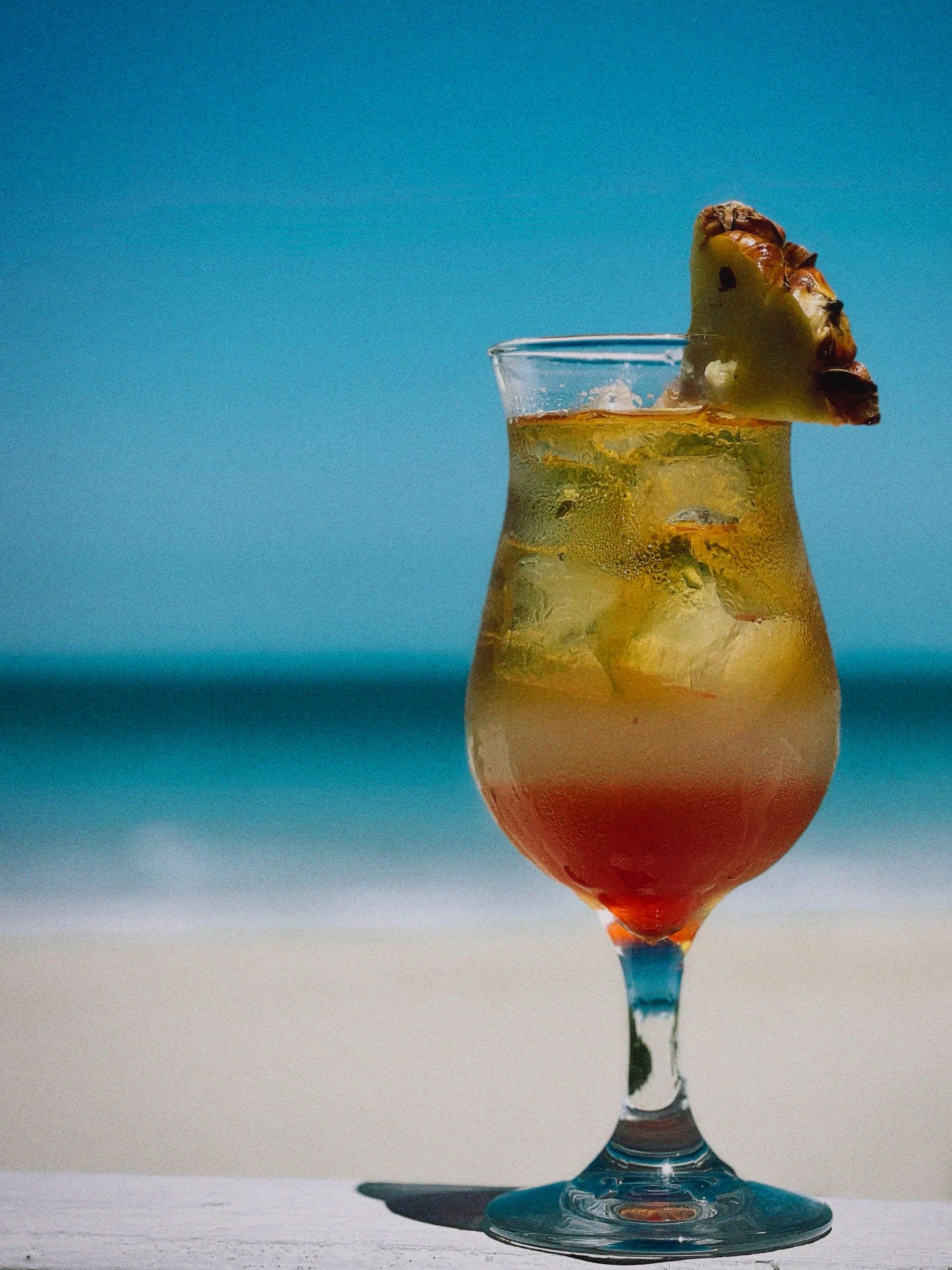 A layered tropical cocktail with ice and a pineapple wedge garnish, sitting on a table with a beach and ocean backdrop.