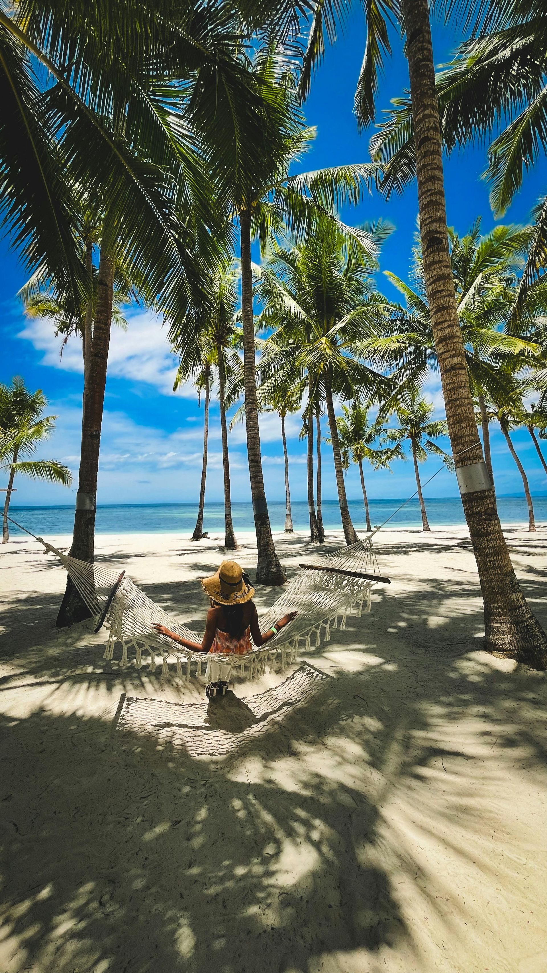Woman in hammock on a sandy beach under palm trees, looking at ocean on a sunny day.