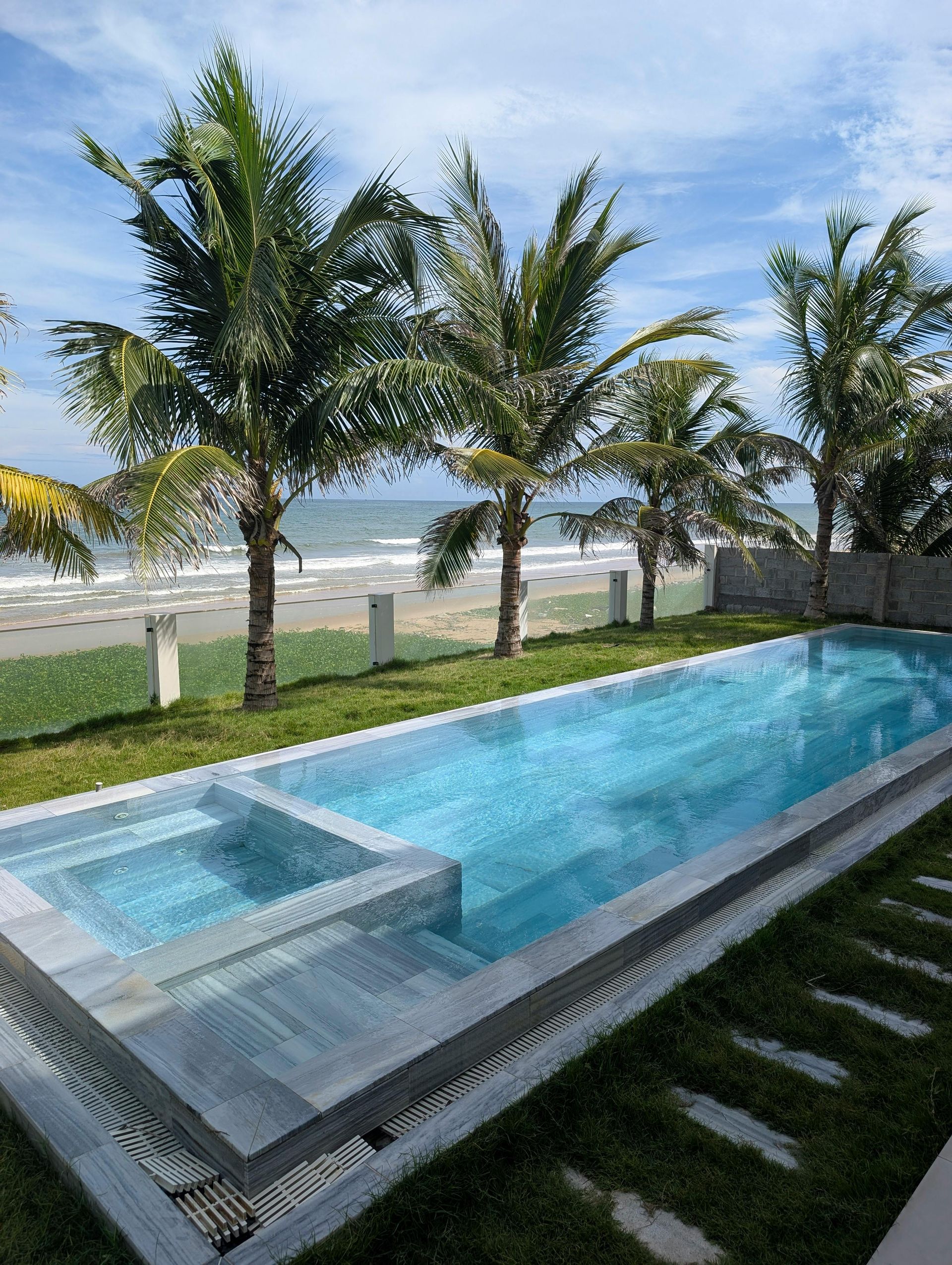 Swimming pool with steps, next to lawn and palm trees, beach and ocean in the background.