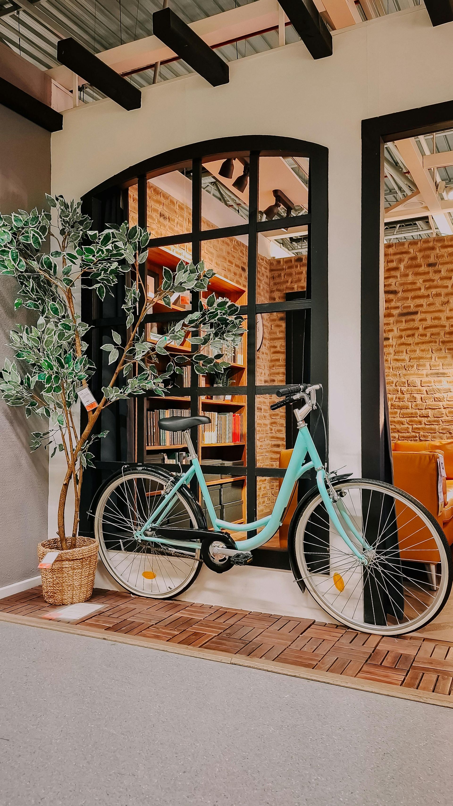 Teal bicycle parked next to a large window. A brick wall and tree are to the left.